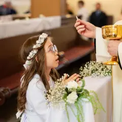 Dziewczynka w wianku na głowie klęczy podczas komunii, przyjmując hostię od księdza.