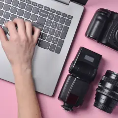 Ręce piszą na laptopie, obok aparat, lampa błyskowa i obiektyw. Szukasz, gdzie sprzedać aparat fotograficzny?