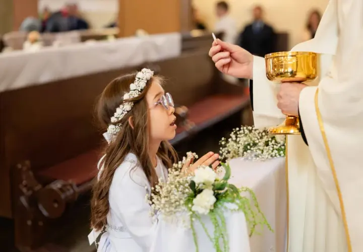 Dziewczynka w wianku na głowie klęczy podczas komunii, przyjmując hostię od księdza.