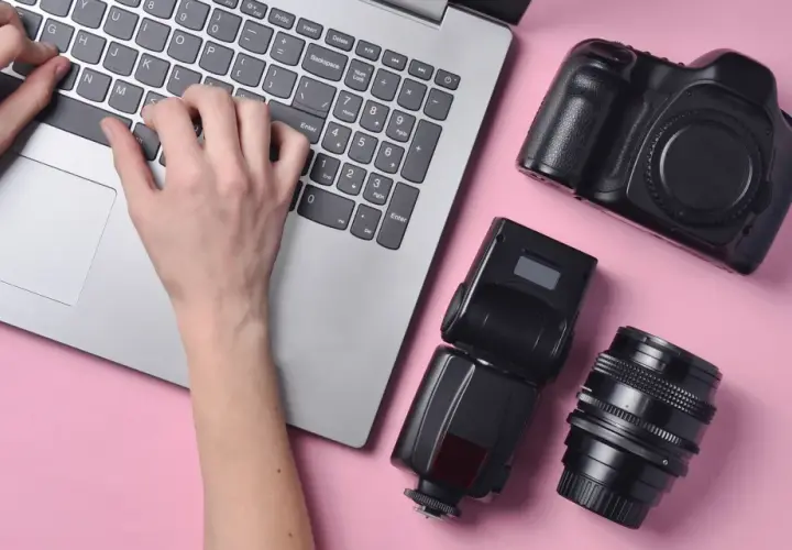 Ręce piszą na laptopie, obok aparat, lampa błyskowa i obiektyw. Szukasz, gdzie sprzedać aparat fotograficzny?