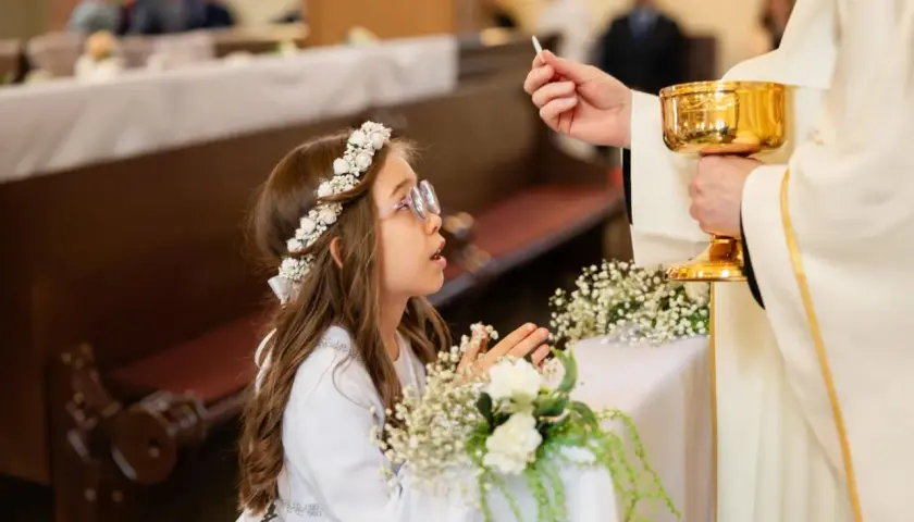 Dziewczynka w wianku na głowie klęczy podczas komunii, przyjmując hostię od księdza.