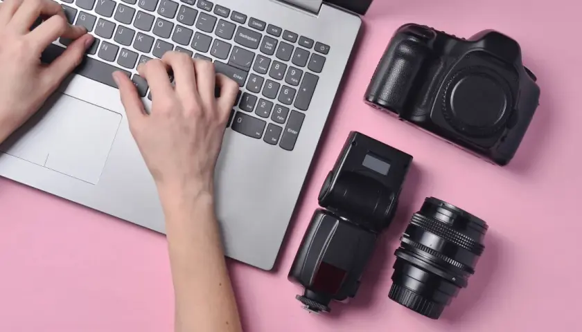 Ręce piszą na laptopie, obok aparat, lampa błyskowa i obiektyw. Szukasz, gdzie sprzedać aparat fotograficzny?