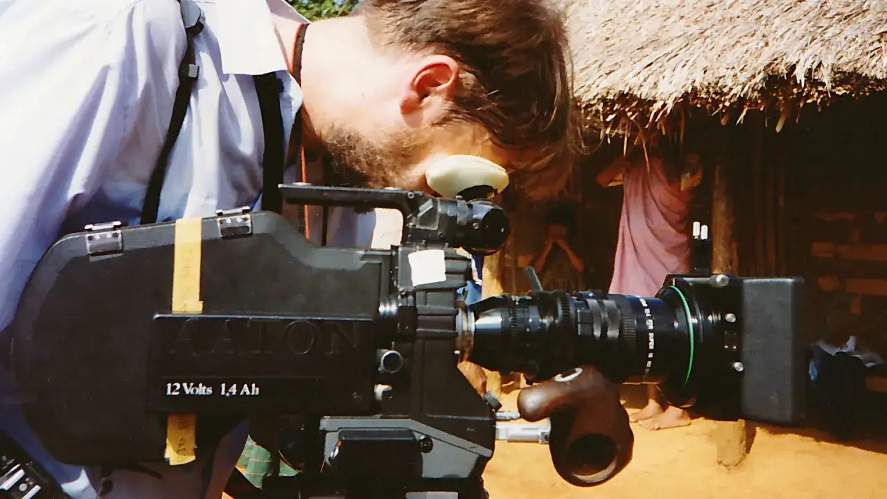 Operator pochyla się nad profesjonalnym aparatem do filmowania Aaton. W tle widać chatę krytą strzechą i ludzi.